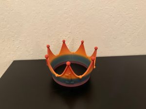 A colorful plastic crown with multiple spikes and round tips sits on a black surface against a textured white wall.